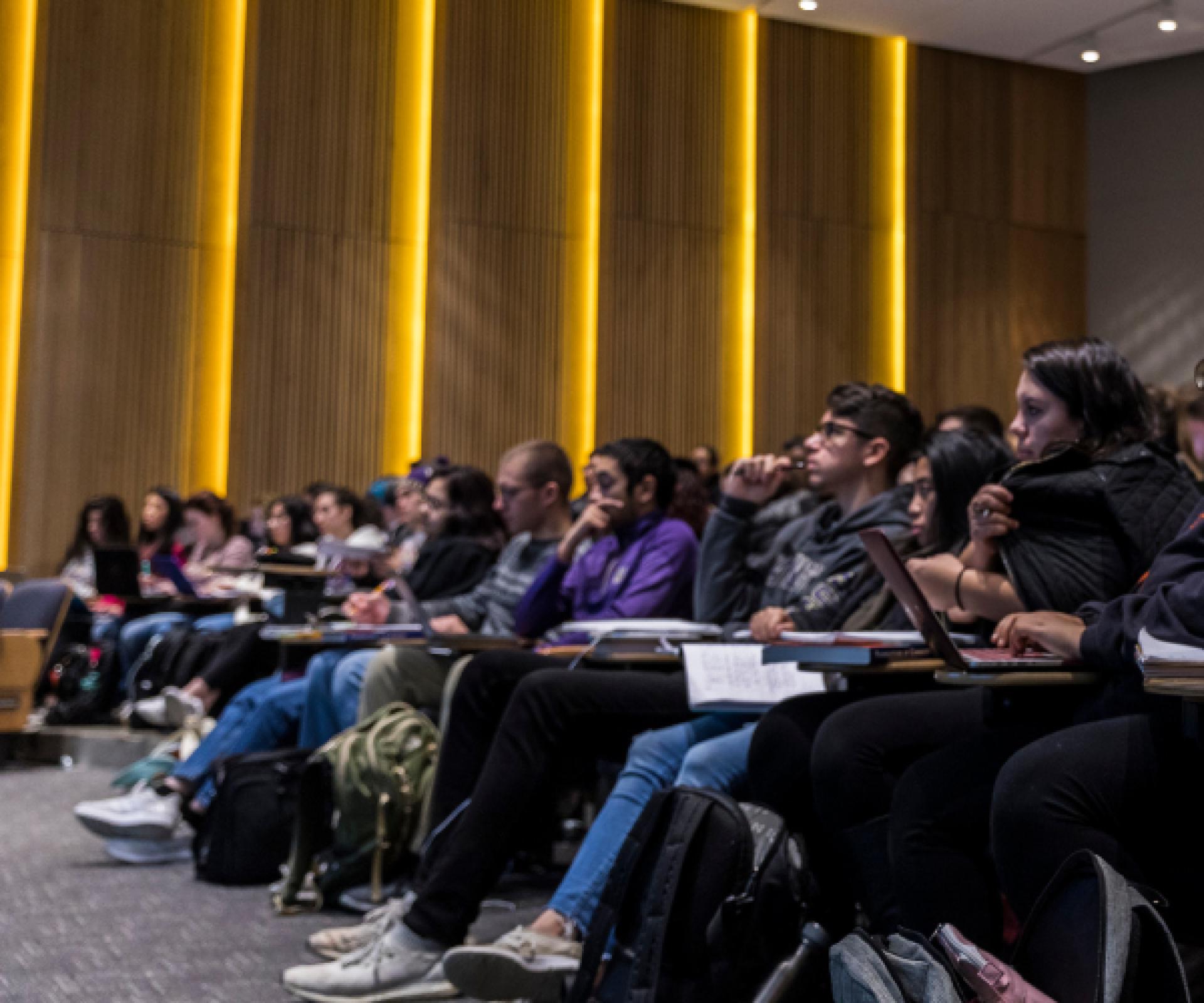 students sitting in the front row of an auditorium like classroom with laptops and notebooks open. There are panels of vertical orange lights behind them.